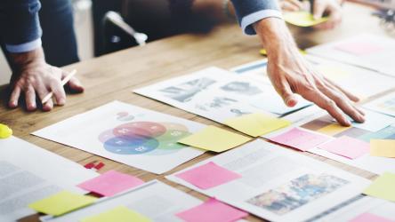 Visuel plusieurs feuilles et post-it posés sur une table en bois avec 2 personnes les organisant