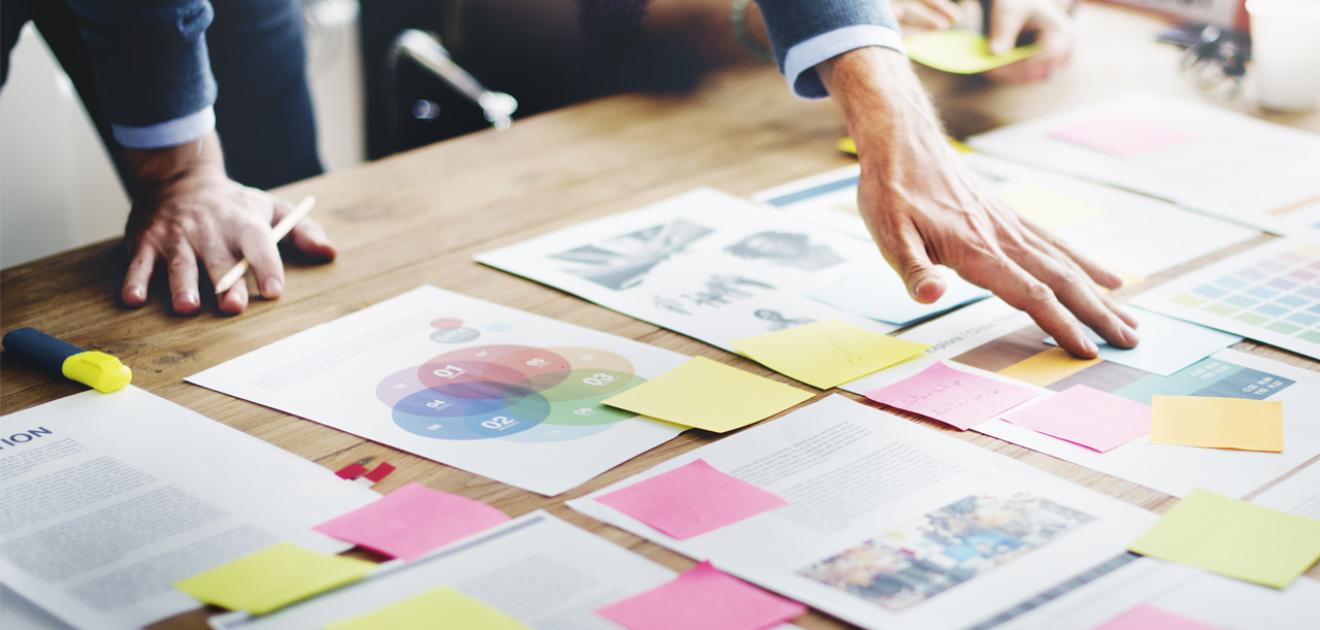 Visuel plusieurs feuilles et post-it posés sur une table en bois avec 2 personnes les organisant